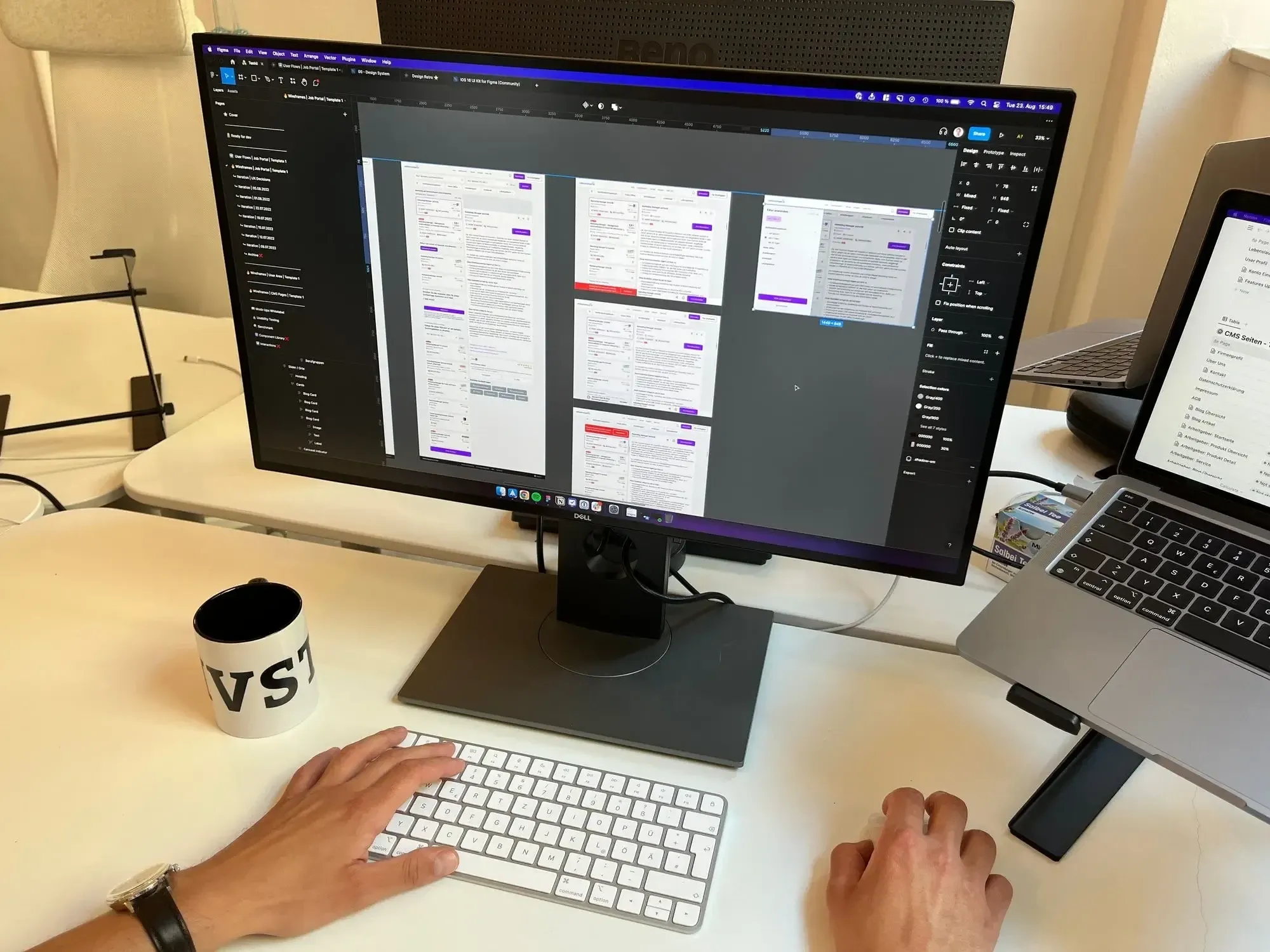 Working desk with white keyboard, white mug, screen showing a Figma file next to a grey laptop.