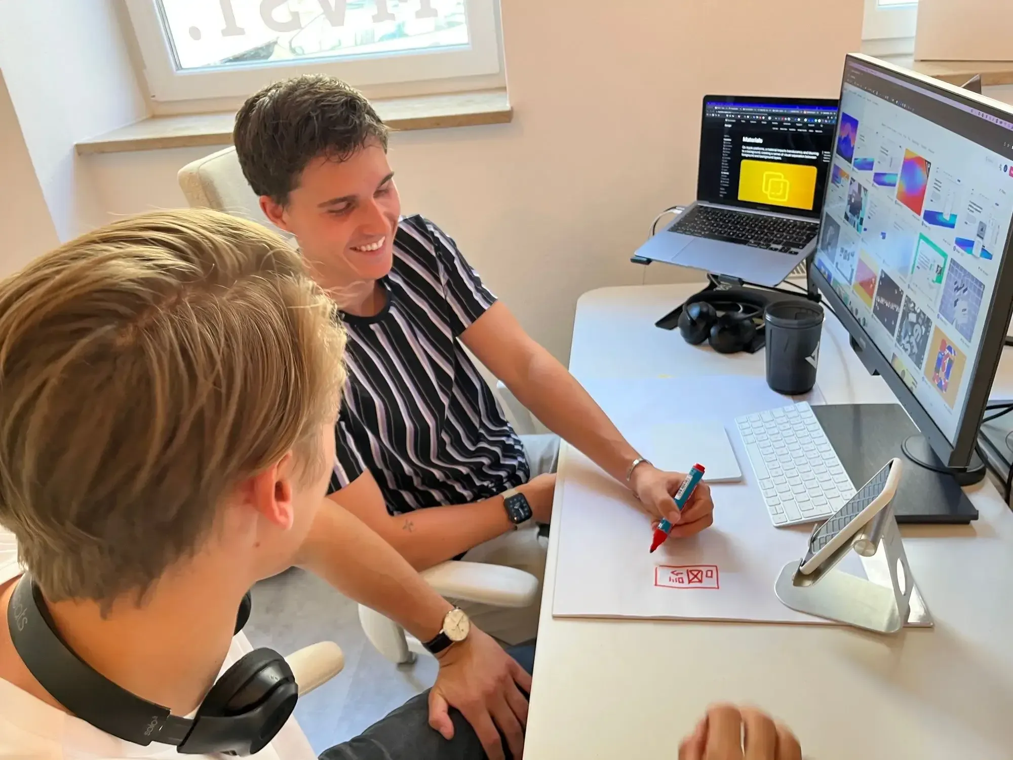 A blond and a brown haired man sitting next to each other in a workspace and discussing designs.